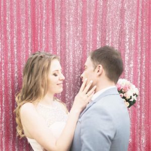 A bride and groom stand together, smiling in front of a shimmering pink sequin backdrop, capturing their joyful moment.
